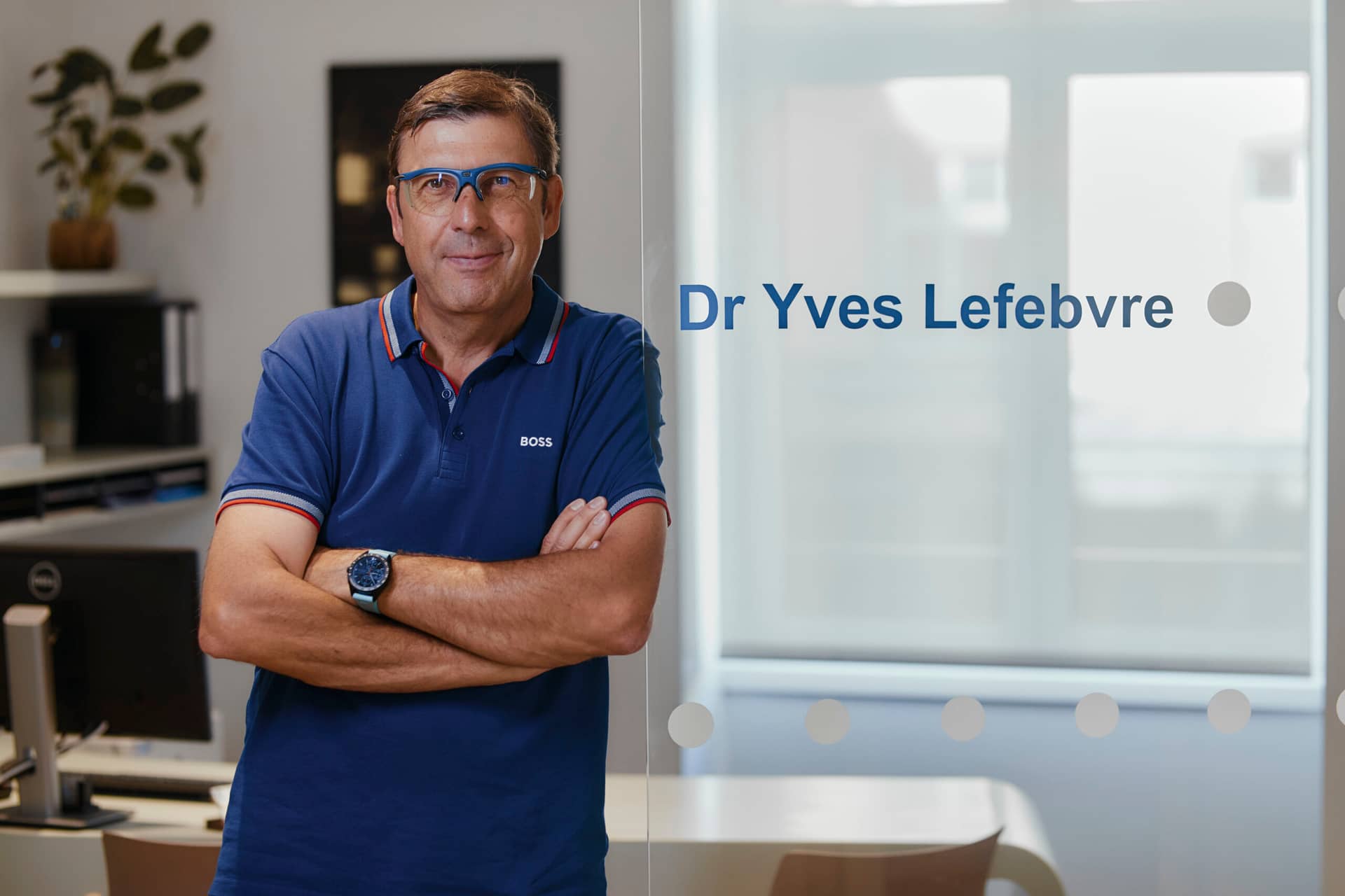 Dr Yves Lefebvre à son bureau à strasbourg Neudorf devant la porte vitrée en polo bleu et lunettes bleues